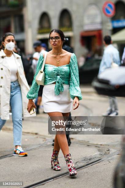 Guest wears glasses, a green shiny sequined square neck long puffy sleeves cropped knotted t-shirt, a white latte leather shoulder bag, a white high...