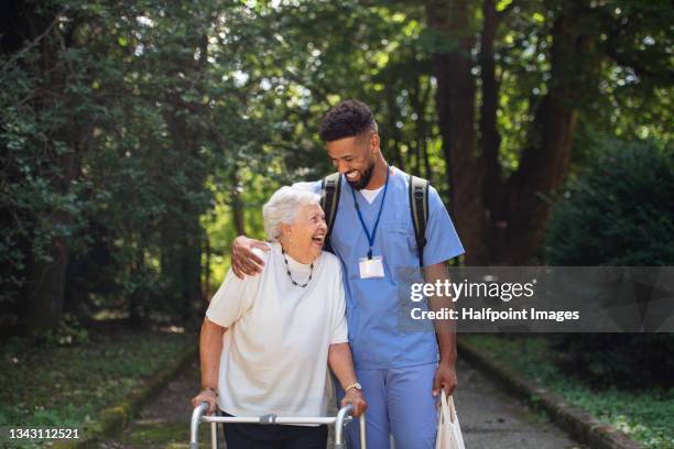 senior woman and caregiver outdoors on a walk with walker in park, talking and laughing - zorgverzekering stockfoto's en -beelden