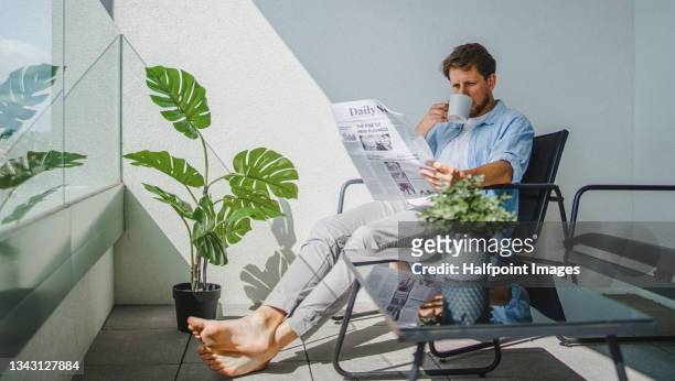 portrait of young man with coffee reading newspaper and sitting outdors on balcony - coffee and news paper foto e immagini stock