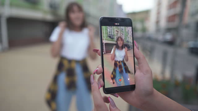 Close-up smartphone screen in female hands with girl dancing having fun on city street. Confident charming Caucasian blogger recording dance for social media outdoors smiling. Vlogging concept.