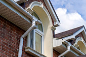 Closeup view of old white gutter system with soffit vent, window with white frame, gutter guard, downspout, decorative trim molding, on corner of brick luxury house