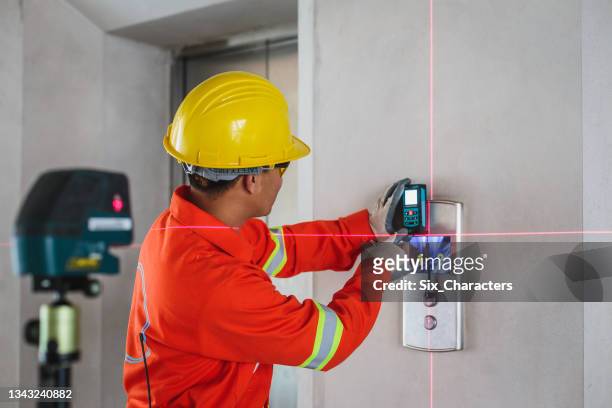 asian construction engineer in hardhat measuring wall with laser leveler next to elevator lift at construction site - elevador stock pictures, royalty-free photos & images
