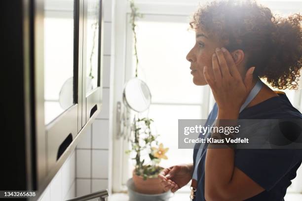 nurse touching face while looking in mirror - mature women skincare stock pictures, royalty-free photos & images