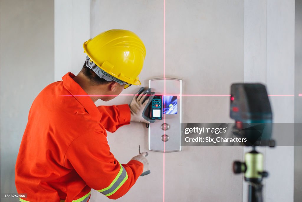 Asian Construction engineer in hardhat measuring wall with laser leveler next to elevator lift at construction site
