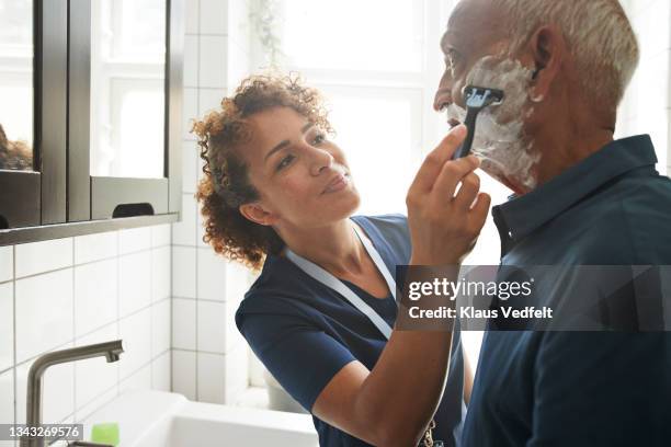 healthcare worker assisting senior man in shaving - asistir fotografías e imágenes de stock