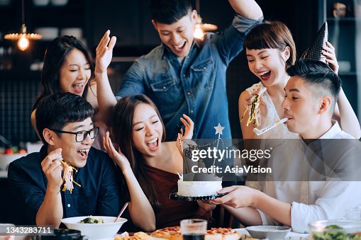 A young group of joyful friends celebrating at a birthday party together. Friends cheering and surprising the birthday girl with a birthday cake during party A young group of joyful friends celebrating at a birthday party together. Friends cheering and surprising the birthday girl with a birthday cake during party