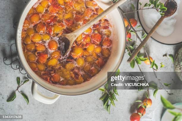 cooked yellow mirabelles in white saucepan with wooden cooking spoon on pale grey concrete kitchen table with branches and green leafs - mirabelle stock-fotos und bilder