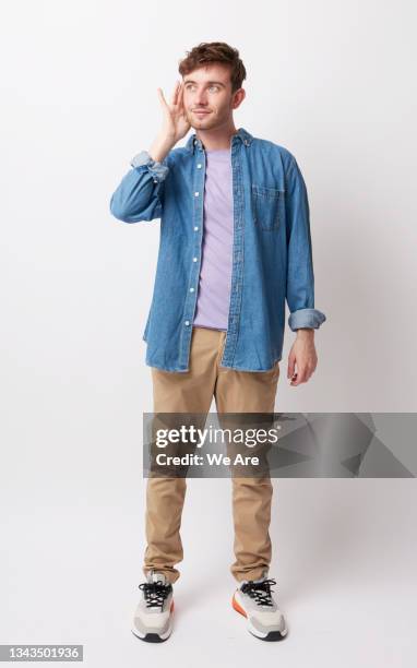 portrait of young man holding hand up to ear listening - junger mann ganzkörper stock-fotos und bilder