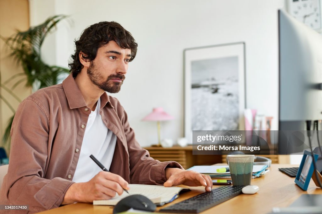 Male Entrepreneur Writing In Book At Home Office