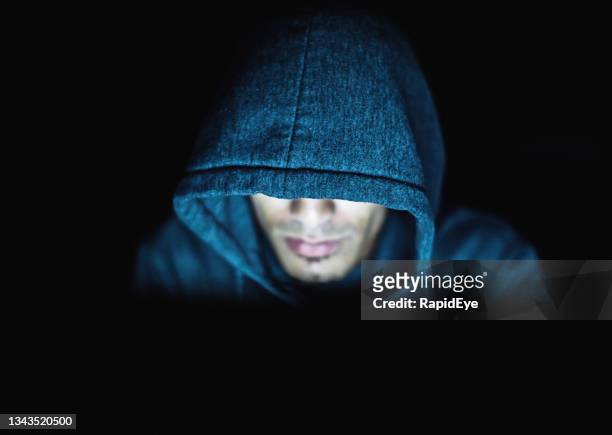 un uomo dall'aspetto inquietante con cappuccio fissa il computer: hacker o cyber-criminale forse - uomo incappucciato foto e immagini stock