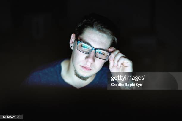 junger mann sitzt am computer, passt seine brille an, arbeitet spät und müde - thick rimmed spectacles stock-fotos und bilder