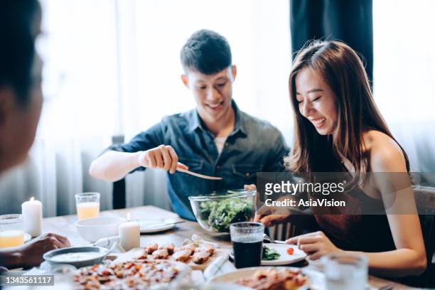 joyful young asian man sharing and serving food to woman while enjoying together with a group of friends. they are having fun, chatting and feasting on food and drinks at dinner party - asian couple dining stockfoto's en -beelden