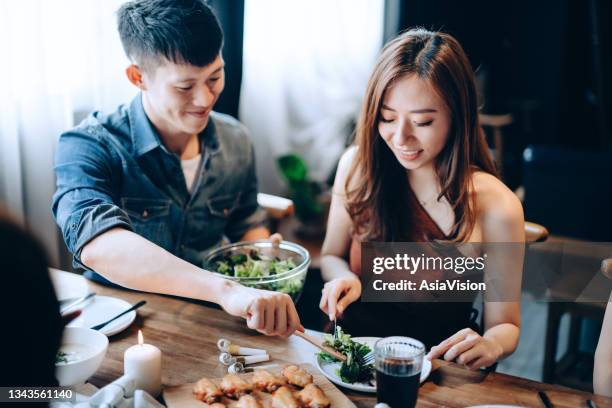 joyful young asian man sharing and serving food to woman while enjoying together with a group of friends. they are having fun, chatting and feasting on food and drinks at dinner party - asian couple dinner stock pictures, royalty-free photos & images