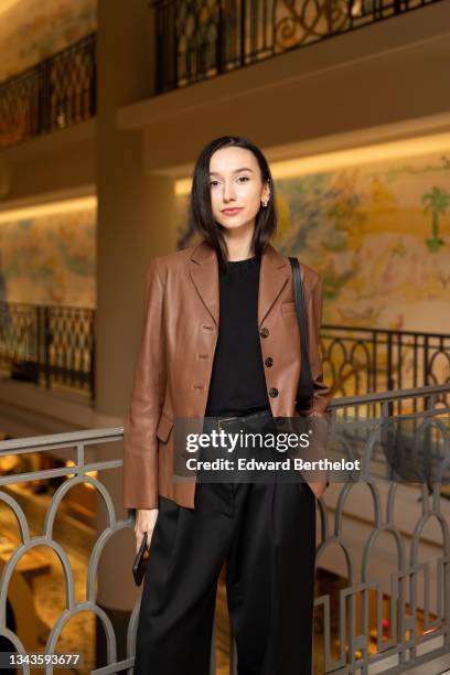 Beatrice Gutu wears a silver ring, a black t-shirt, a brown shiny leather blazer jacket, a black shiny leather belt, high waist black flowing / large...