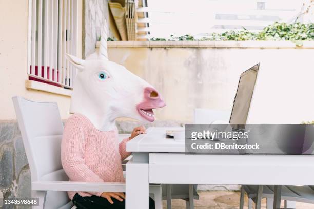 child wearing a unicorn costume latex mask, wearing a pink sweater, sitting in front of the laptop, on the terrace. online distance education, childhood, technology and surrealism concept. - cow head fotografías e imágenes de stock