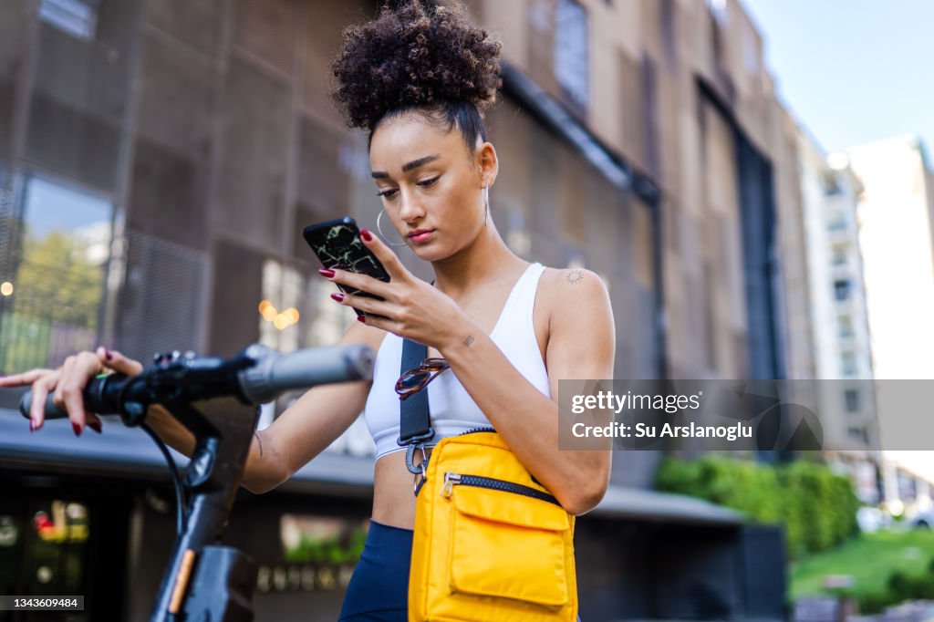 Mixed race young girl standing on the street with electric scooter while using smart phone