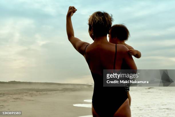 the back view of a woman standing on the beach, holding a baby in one hand and raising the other arm high. - braveheart imagens e fotografias de stock