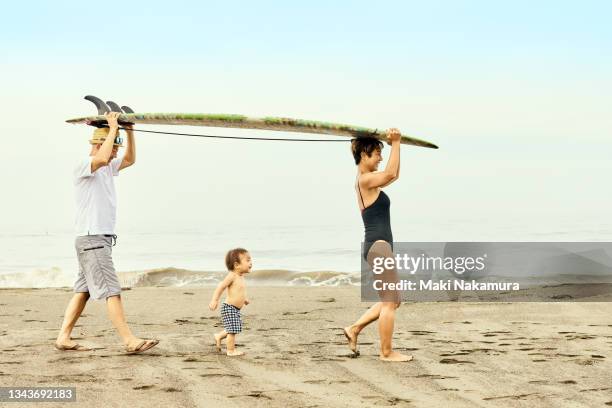 a child under the age of one is walking in the middle of a couple carrying a surfboard on their heads on the beach. - baby lachen natur stock-fotos und bilder