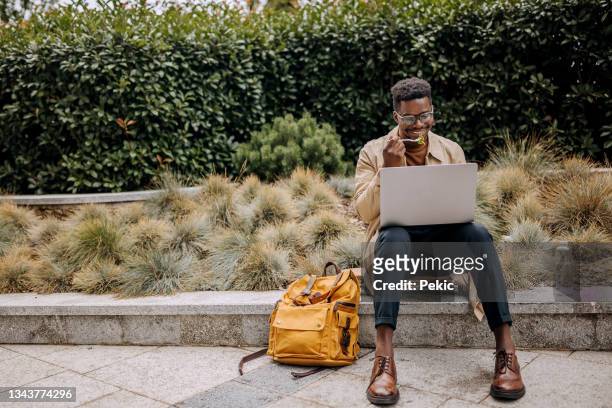 gutaussehender junger geschäftsmann beim mittagessen, während er den laptop im öffentlichen park benutzt - snacking on the go stock-fotos und bilder