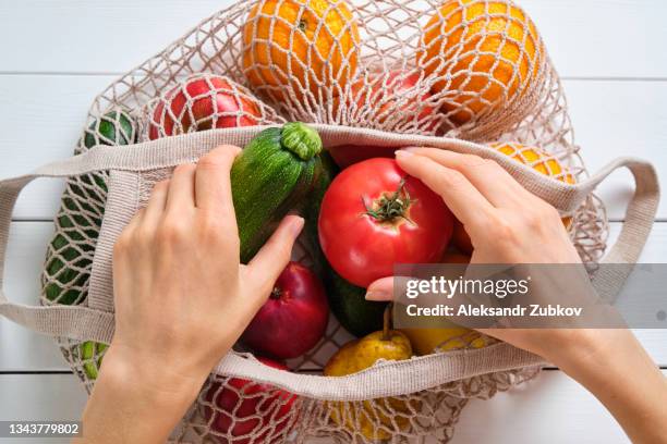 fresh juicy fruits and vegetables in a reusable shopping bag. a girl or a woman takes or lays out products from a string bag made of recycled materials on a white wooden table or background. vegetarianism, veganism, raw food. online shopping. no plastic. - plastic free stock pictures, royalty-free photos & images