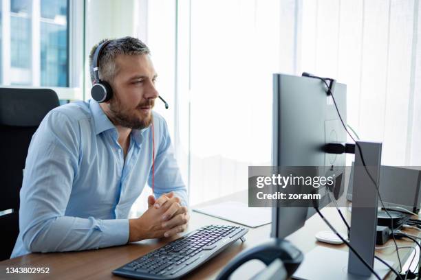 mann mit kopfhörern sitzt am schreibtisch, ausgestattet mit computer - headset stock-fotos und bilder