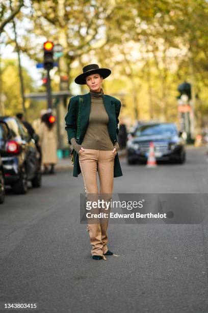 Landiana Cerciu wears a black with black strap hat, a gold turtleneck pullover, a green blazer jacket, a green with orange / yellow painted pattern...