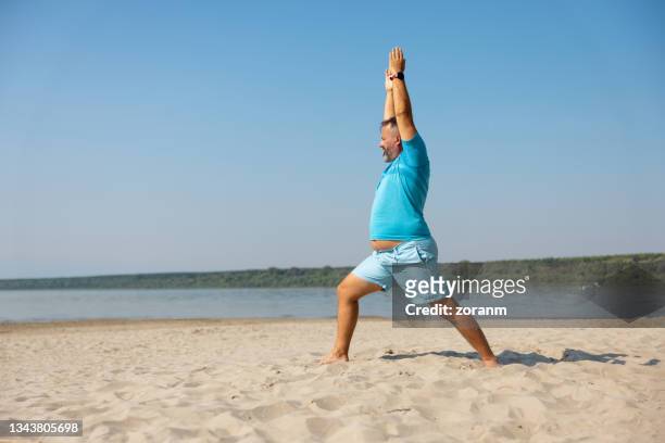 homme pratiquant la posture de yoga sur la plage par temps clair - fat guy on beach photos et images de collection