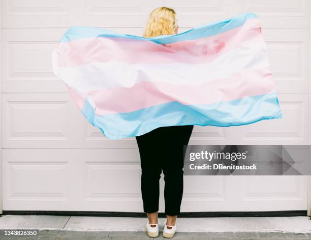 transgender person from behind, wearing pink and white striped sweatshirt, holds transgender flag - transessuale foto e immagini stock