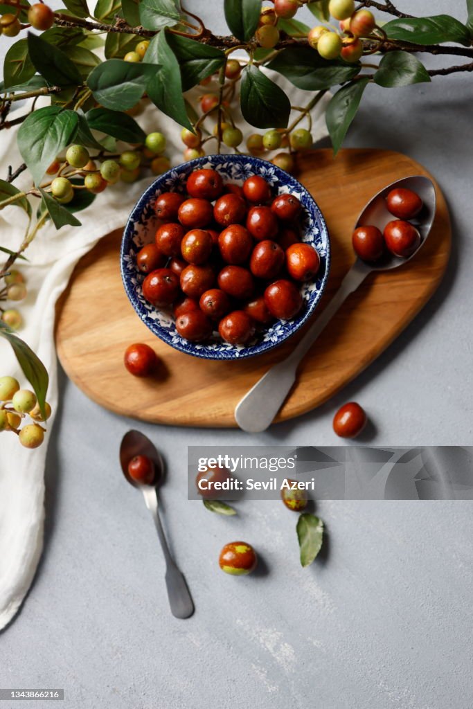 Fresh small jujubes in a rustic ceramic bowl with a blue floral pattern on a light gray rough concrete floor. Around the jujube bowl, there are tree branches with green-yellow berries and green leaves. Hünnap.