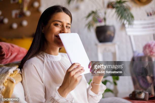 cheerful young woman hiding her smile behind a love letter from a crush - love letter stock pictures, royalty-free photos & images