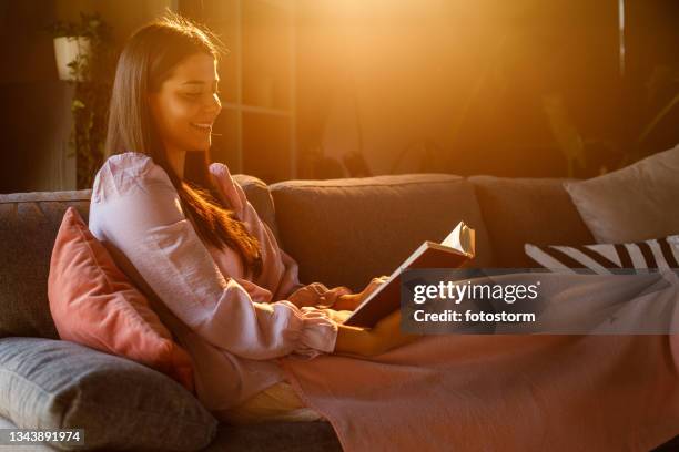 young woman relaxing on the sofa under a cozy blanket, reading a book - zacht licht stockfoto's en -beelden