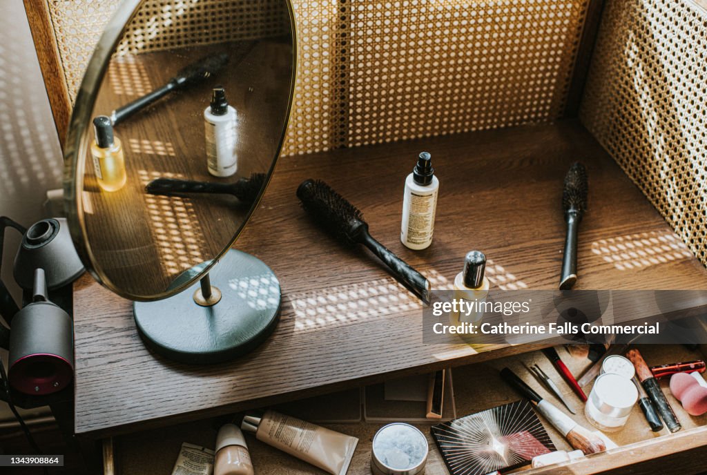 A wooden dressing table in sun with various beauty products, including a hairdryer and diffuser