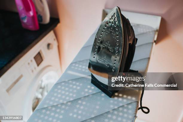 an iron sitting upright on an ironing board, with a washing machine visible in the background. - iron fotografías e imágenes de stock