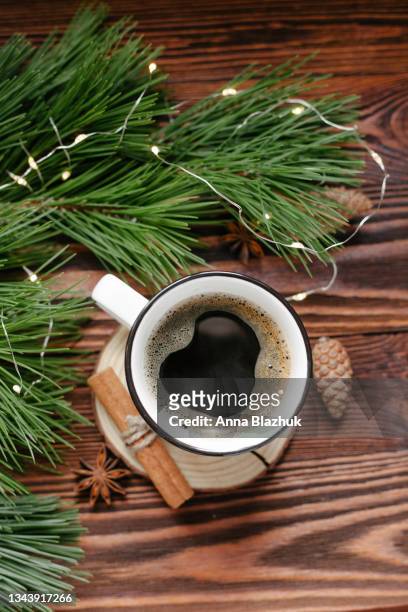 white cup of hot coffee. christmas greeting card. natural pine tree green branches, winter decorations of pine cones and fairy lights over dark brown wood background. - coffee christmas ストックフォトと画像