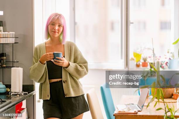 porträt einer jungen frau, die eine tasse heißes getränk hält und in der küche zu hause vor die kamera lächelt - woman portrait kitchen laptop stock-fotos und bilder