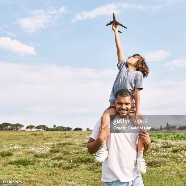 a young boy with a toy aeroplane being carried on his fathers shoulders in a meadow. - on shoulders stock pictures, royalty-free photos & images