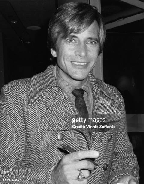 American actor Dirk Benedict wearing a tweed jacket over a checked shirt and tie, smiling with a pen in his right hand, location unspecified, circa...