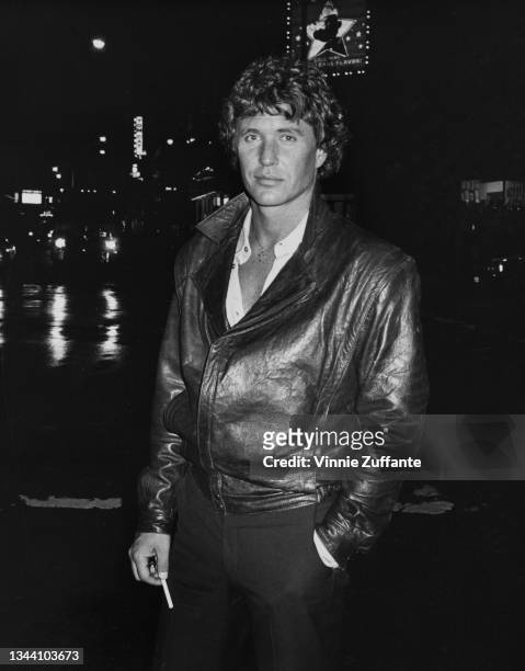 American actor Tom Berenger, wearing a leather jacket, stands on an unspecified street by night with illuminated signs in the background, on the set...