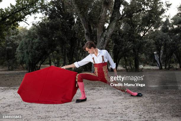 side view of slim male bullfighter in red pants and white shirt holding cloak performing gracefully before - bullfight photos et images de collection