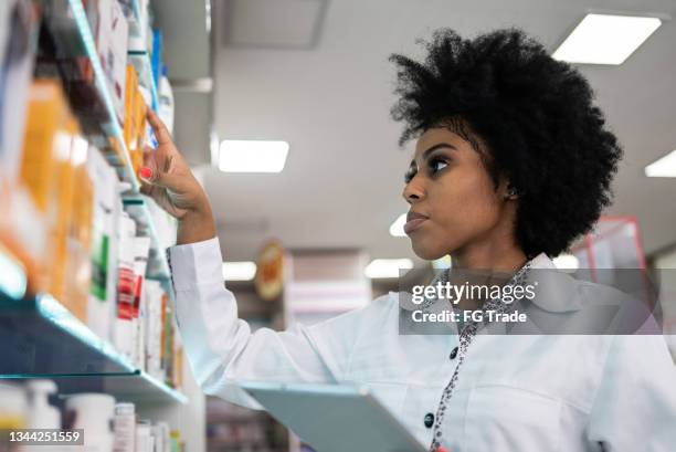 young pharmacist checking the shelves with a digital tablet at the pharmacy - female pharmacist with a digital tablet imagens e fotografias de stock