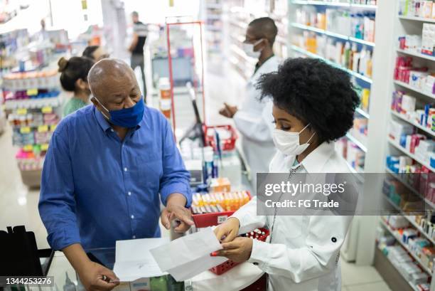 junger apotheker hilft einem älteren mann in der apotheke - mit gesichtsmaske - pharmacist stock-fotos und bilder