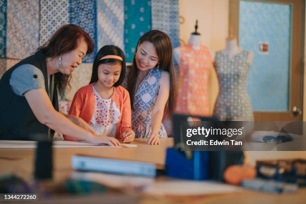 asiatische chinesische großmutter aus mehreren generationen, die ihrer enkelin das zeichnen von kleidungsdesign in ihrem atelier beibringt - modeatelier stock-fotos und bilder
