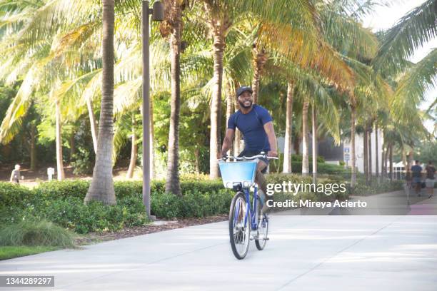 an african american millennial man with beard rides a bike through a tree lined sidewalk through the city, wearing athletic clothing. - miami fahrrad stock-fotos und bilder