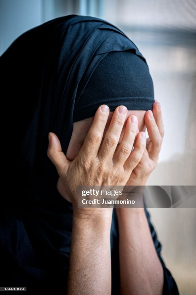 Sad muslim woman looking through a window covering her face with her hands