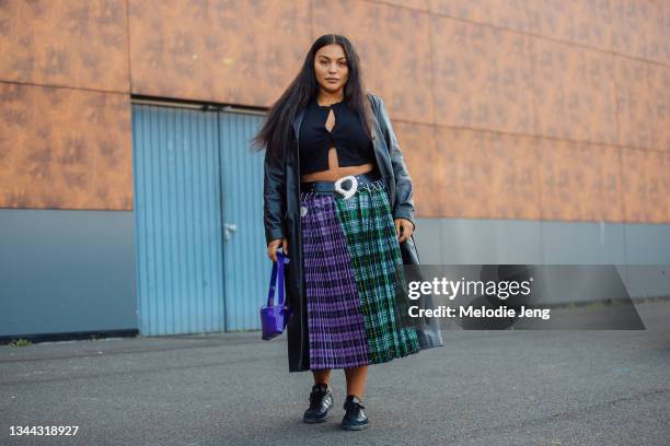 Model Paloma Elsesser wears a black trench coat, black croptop, green and purple plaid print skirt, blue bag, and black Adidas sneakers at the...