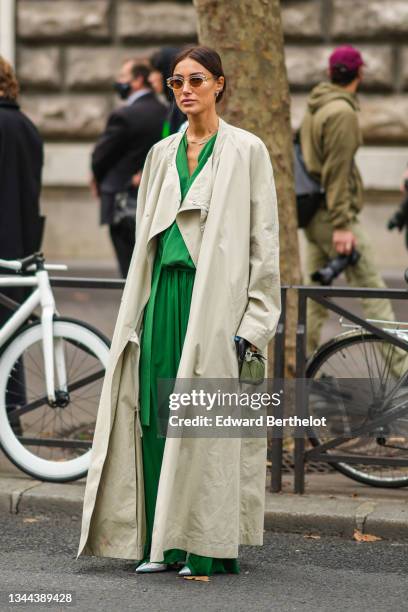 Giorgia Tordini wears sunglasses, earrings, a green V-neck flowing oversized belted jumpsuit, a beige long oversized coat, gold chain necklaces,...