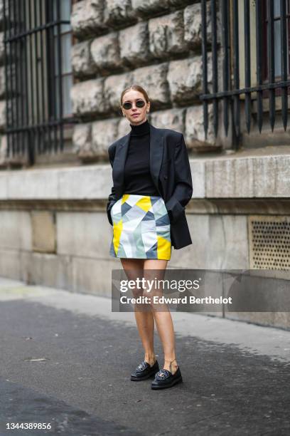 Pernille Teisbaek wears sunglasses, earrings, a black turtleneck pullover, a black long blazer jacket, a yellow / gray / black / green / white print...