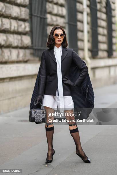 Miss Fame wears sunglasses, a white long shirt dress, a black blazer jacket with a black ribbed embroidered cape / cloak in the back from Loewe,...
