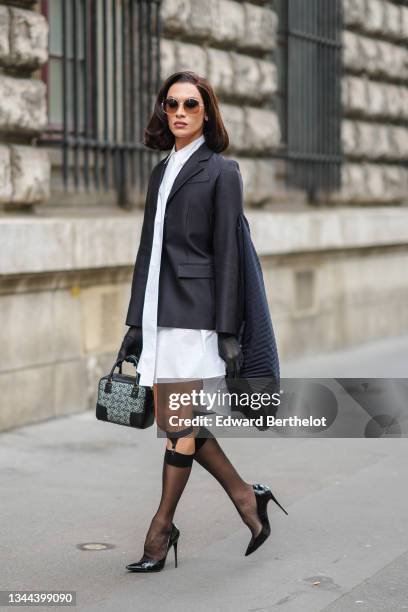 Miss Fame wears sunglasses, a white long shirt dress, a black blazer jacket with a black ribbed embroidered cape / cloak in the back from Loewe,...