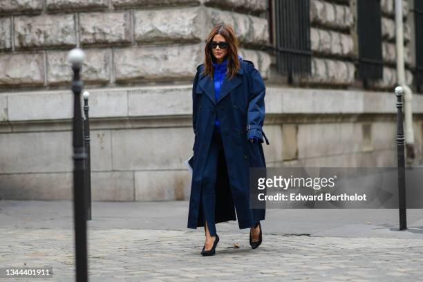 Christine Centenera wears black sunglasses, a royal blue electric hoodie sweater, a navy blue oversized long coat, navy blue slit / split legging...